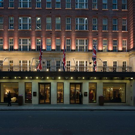 The May Fair, A Radisson Collection Hotel, Mayfair London Exterior photo The photo depicts a grand hotel exterior at dusk. The building has a classic architectural style, characterized by multiple stories with large windows. The facade is illuminated, highlighting the architectural features. British flags are prominently 