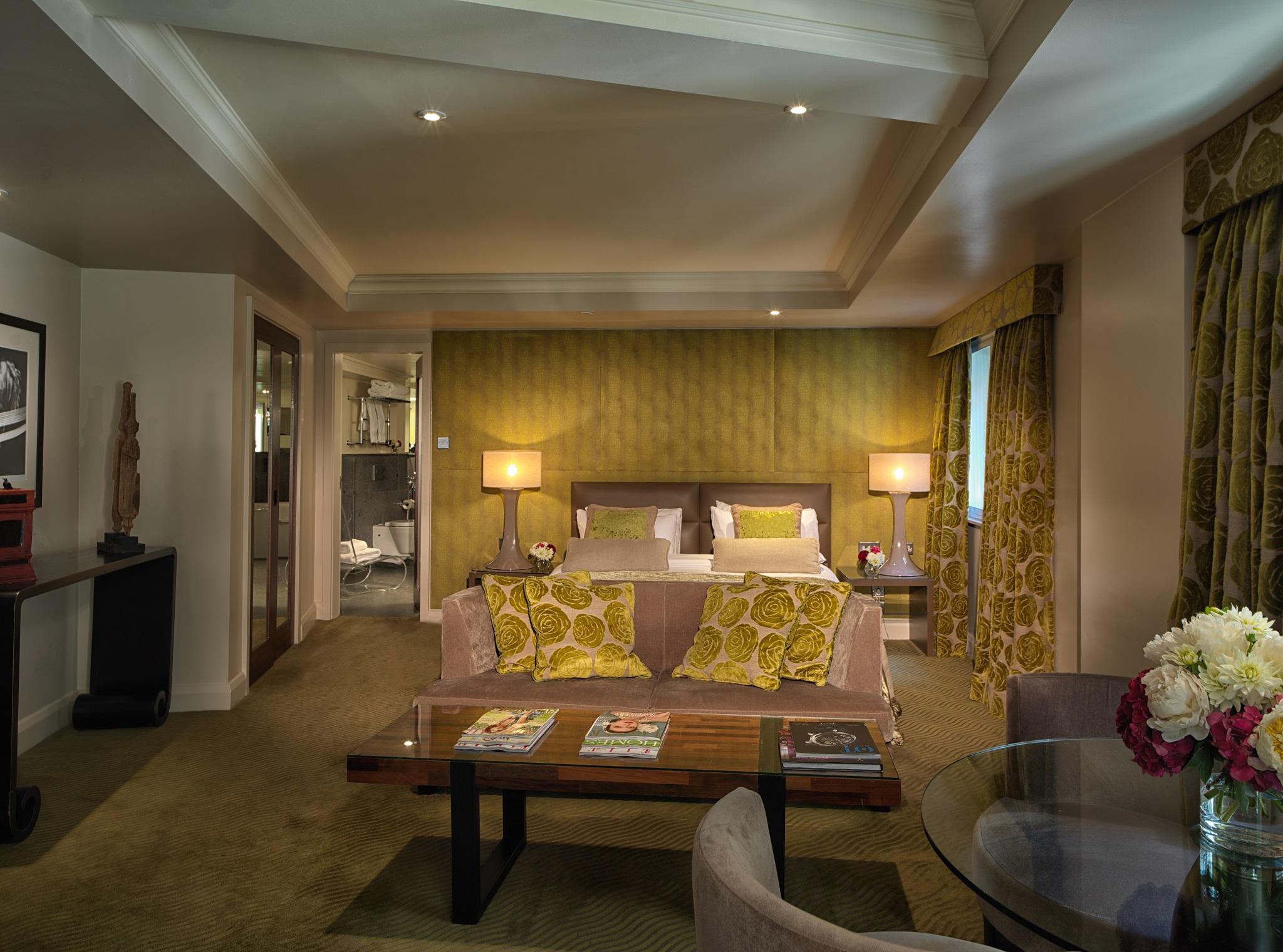 The May Fair, A Radisson Collection Hotel, Mayfair London Exterior photo The photo showcases a stylish hotel room or suite. It features a seating area with a couch adorned with decorative yellow pillows and a coffee table. There are two lamps on either side of the couch providing soft lighting. The walls are painted in a 