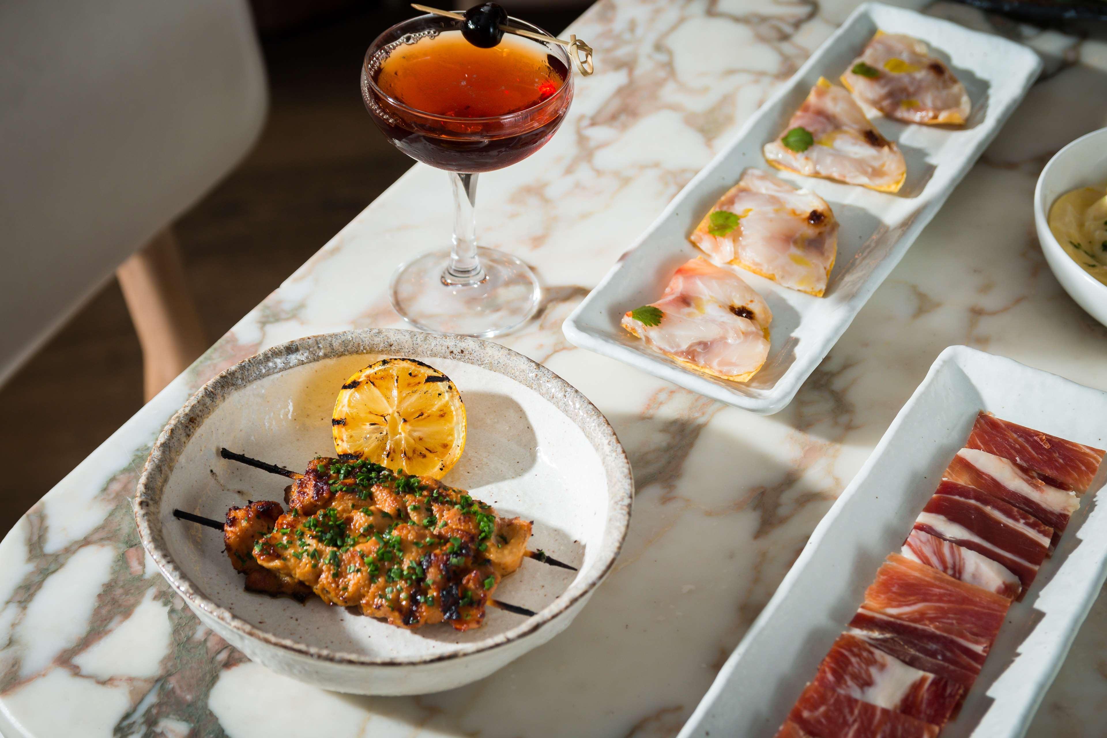 The May Fair, A Radisson Collection Hotel, Mayfair London Exterior photo The photo features a beautifully arranged dining table with a variety of food and a drink. In the foreground, there is a dish containing grilled or cooked meat, garnished with herbs and served with a roasted lemon slice. Next to it, a cocktail drink 