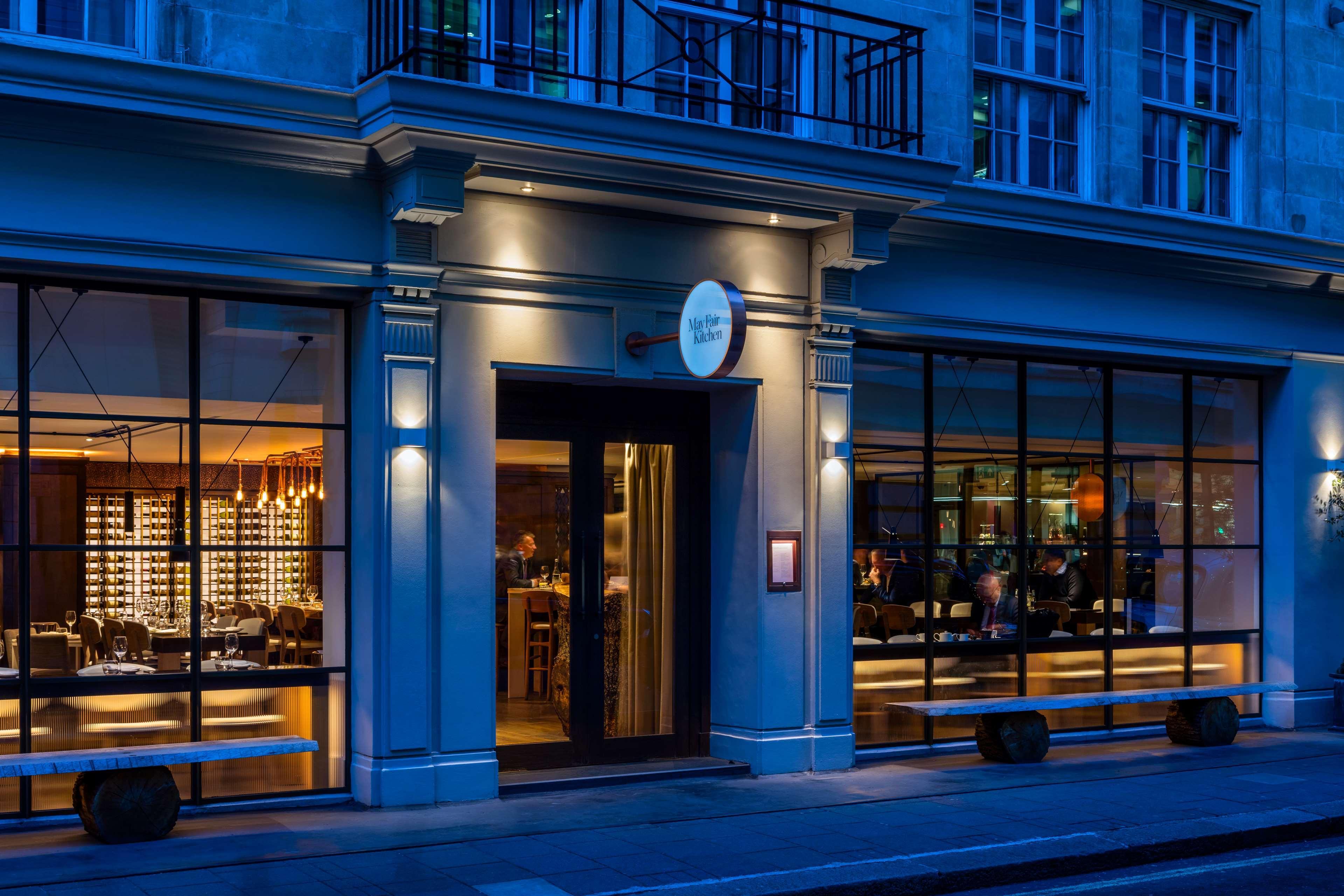 The May Fair, A Radisson Collection Hotel, Mayfair London Exterior photo The photo shows the exterior of a stylish restaurant. The entrance features a glass door and large windows, allowing a view inside. The restaurant is illuminated with soft lighting, creating a warm and inviting atmosphere against the evening backdrop