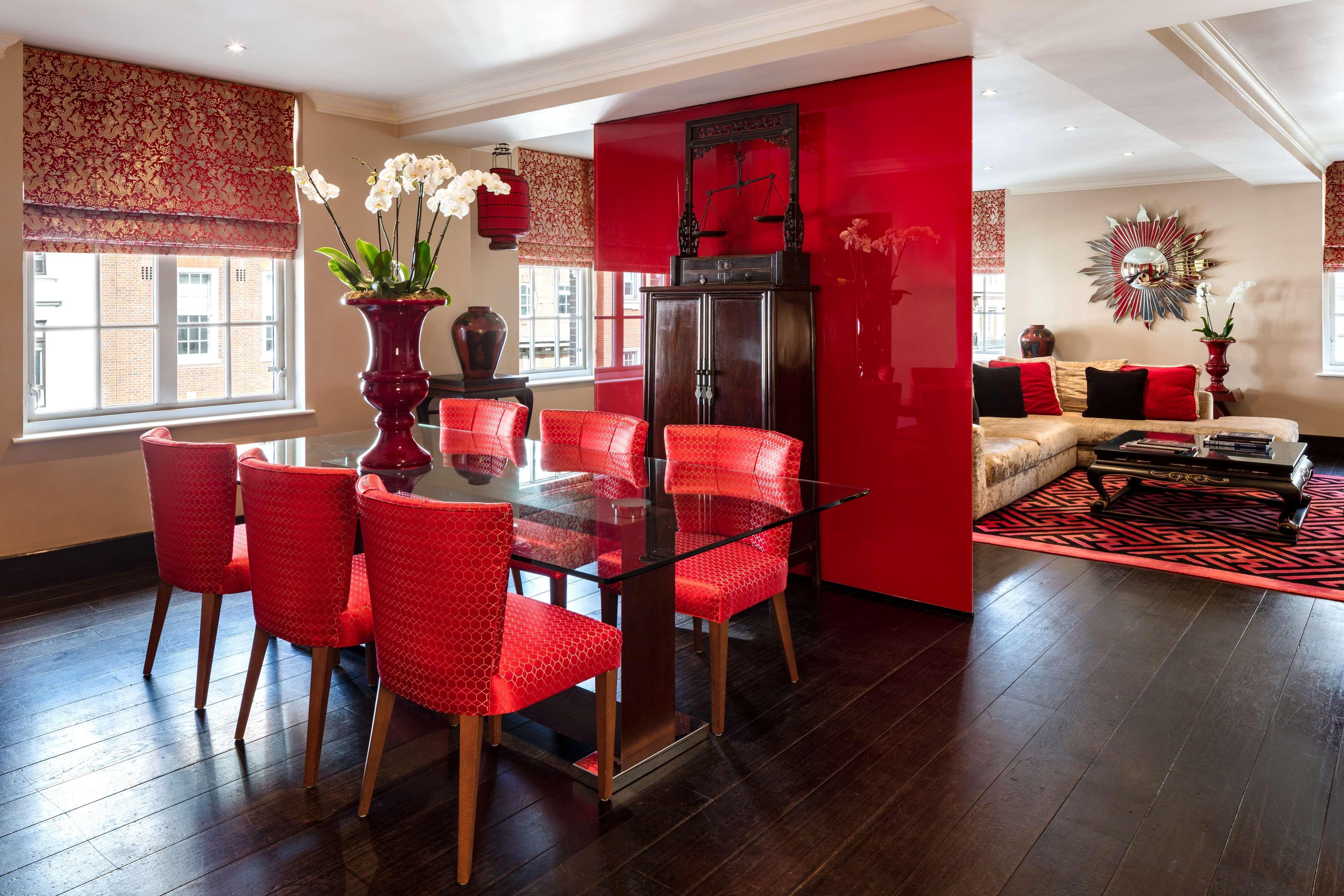 The May Fair, A Radisson Collection Hotel, Mayfair London Exterior photo The photo depicts a stylish, modern living and dining area. 

- **Dining Area**: There is a glass dining table with six vibrant red chairs surrounding it. A large vase containing white orchids sits at the center of the table.
  
- **Decor Elements**: