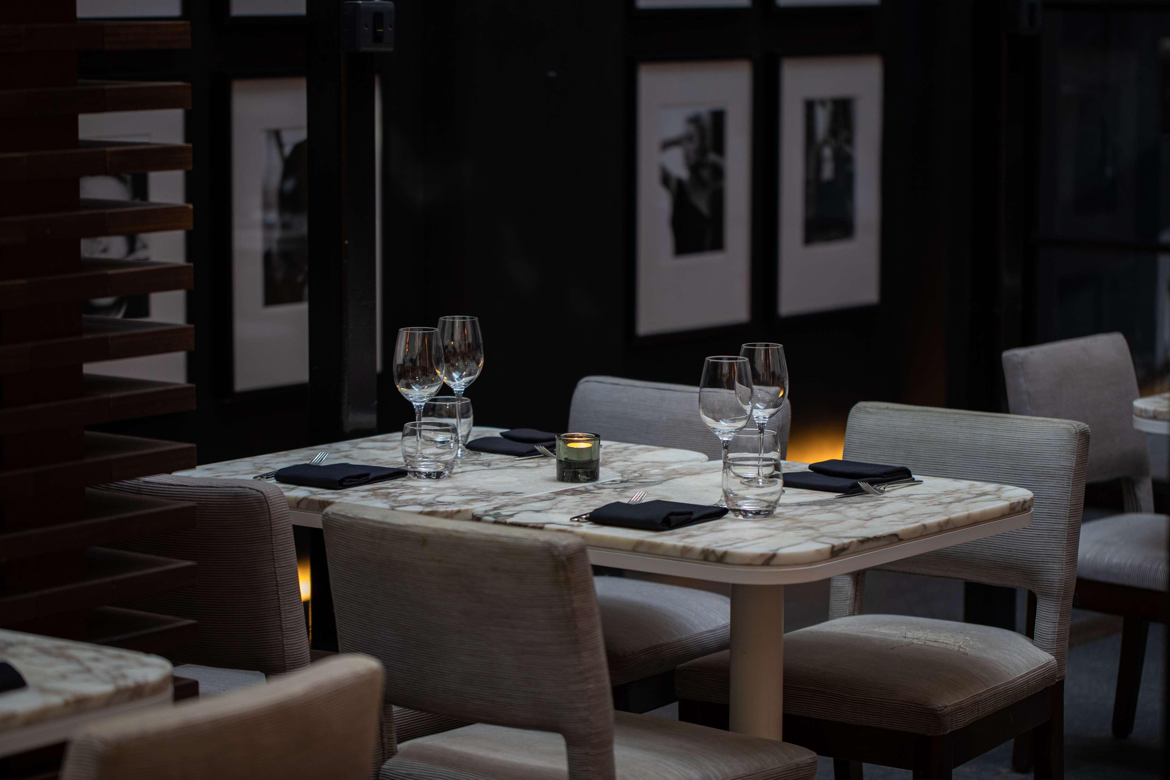 The May Fair, A Radisson Collection Hotel, Mayfair London Exterior photo The photo shows a dining area with a stylish and elegant atmosphere. There are two tables set for dining, featuring marble tops and light-colored chairs. Each table has wine glasses and a small candle holder, creating a warm ambiance. In the backgrou