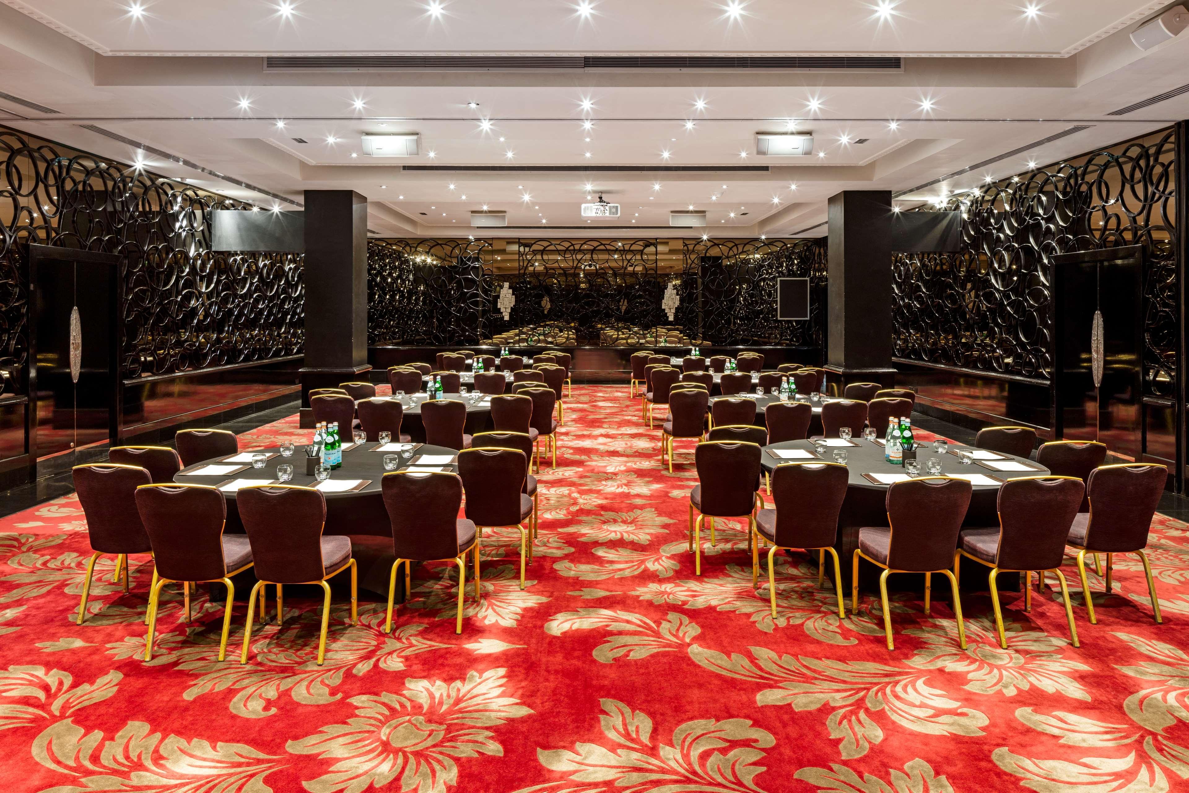 The May Fair, A Radisson Collection Hotel, Mayfair London Exterior photo The photo shows a spacious dining or banquet room. It features several round tables arranged neatly with dark chairs. The floor is covered with a vibrant red carpet that has floral patterns. The walls are decorated with a sleek, dark design, adding a