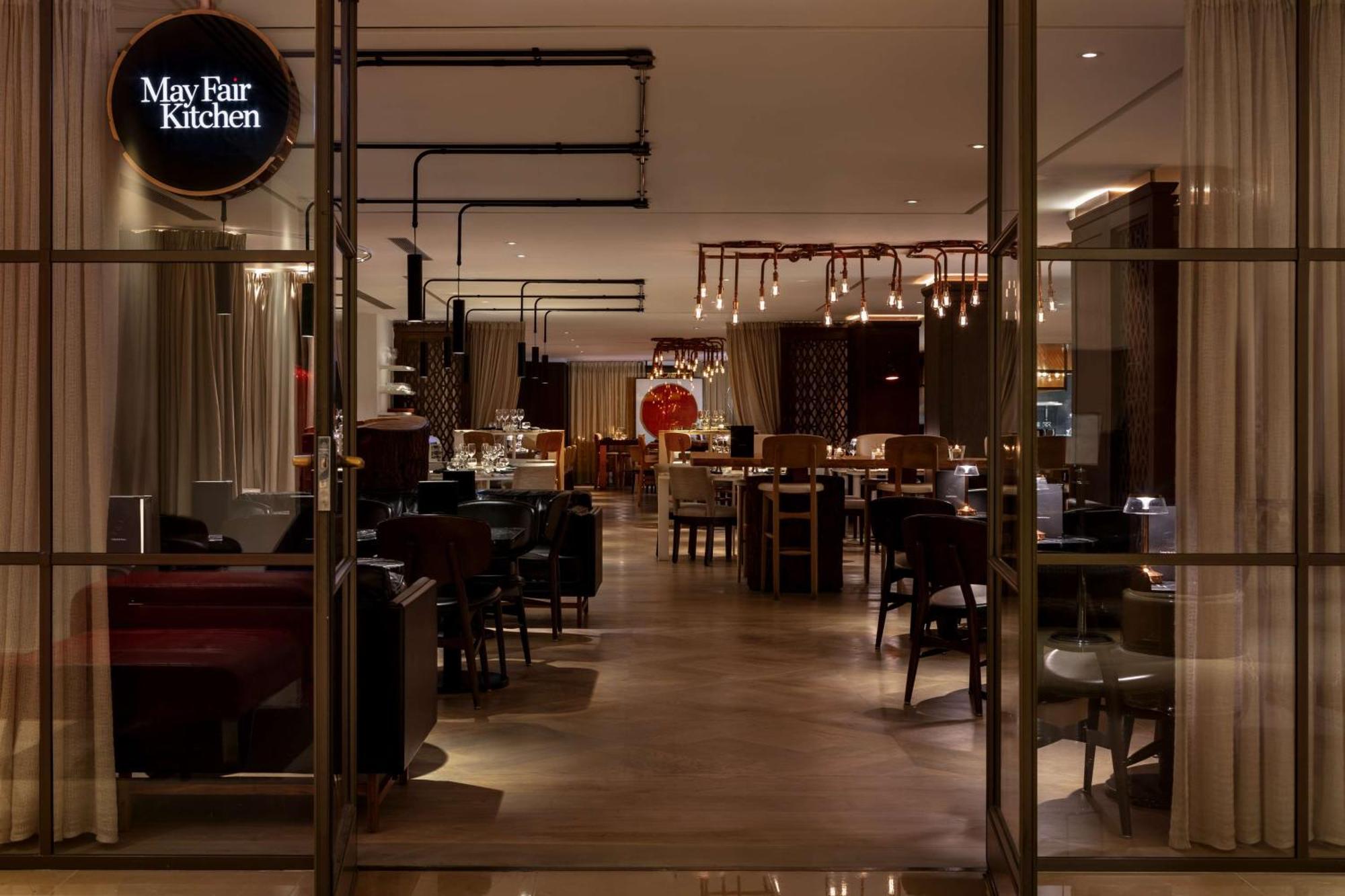 The May Fair, A Radisson Collection Hotel, Mayfair London Exterior photo The photo shows the interior of a restaurant called "May Fair Kitchen." The setting features a modern and elegant design, with various tables and chairs arranged throughout the space. There are warm lighting fixtures hanging from the ceiling, and the