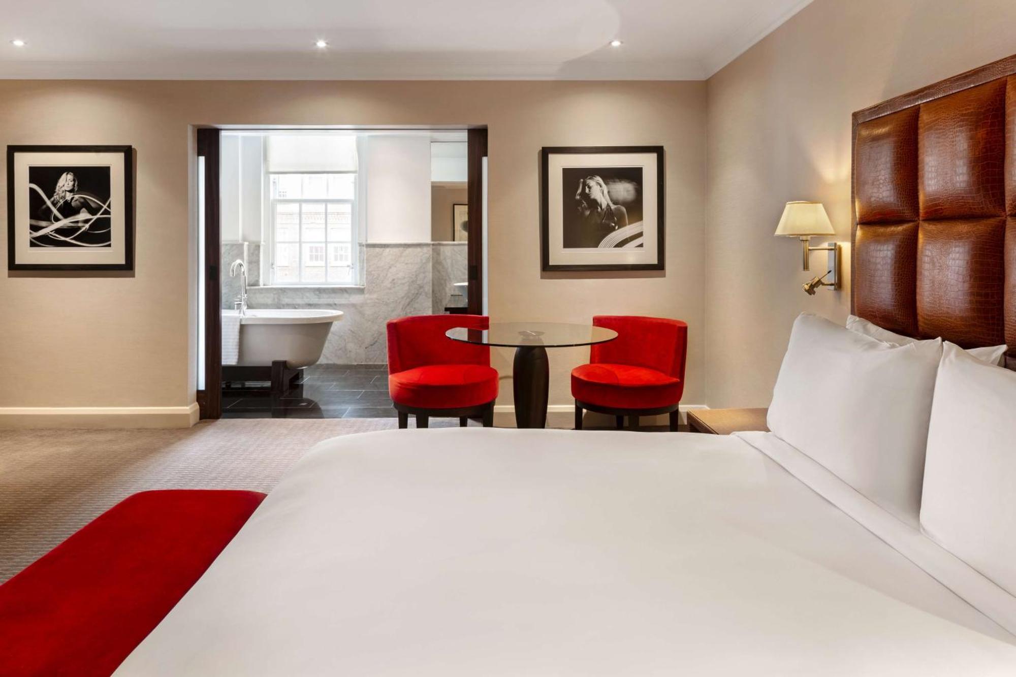 The May Fair, A Radisson Collection Hotel, Mayfair London Exterior photo The photo shows a hotel room featuring a large bed with white linens and a red accent throw. In the background, there are two modern red chairs around a small glass table. The wall displays framed black and white photographs. To the side, there is an