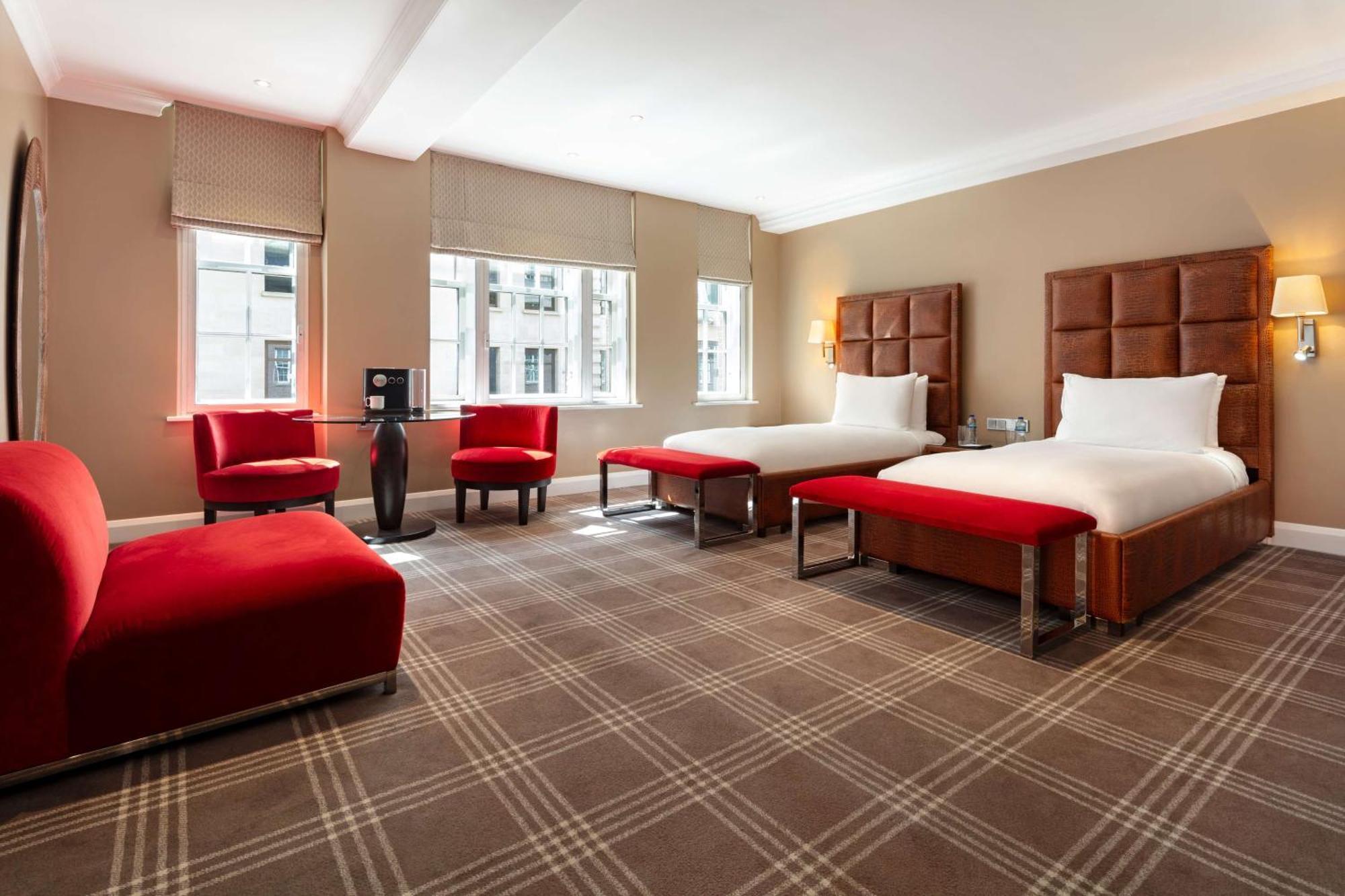 The May Fair, A Radisson Collection Hotel, Mayfair London Exterior photo The photo shows a modern hotel room featuring two beds with white bedding and brown headboards. There are two red benches at the foot of each bed. A red upholstered chair and a small round table are positioned near the window, which allows natural li