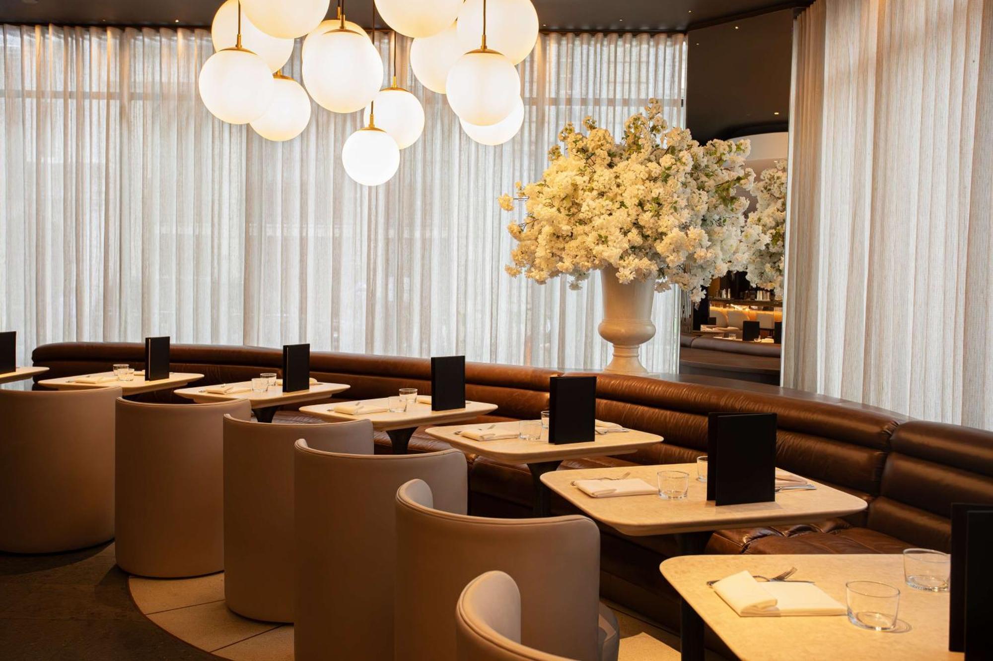 The May Fair, A Radisson Collection Hotel, Mayfair London Exterior photo The photo shows a modern restaurant interior. It features stylish seating arrangements with curved booths and elegant tables. The lighting is soft, provided by large, round pendant lights hanging from the ceiling. A large floral arrangement in a whit